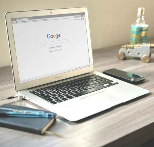 A neat workspace featuring a laptop displaying Google search, a smartphone, and a notebook on a wooden desk.